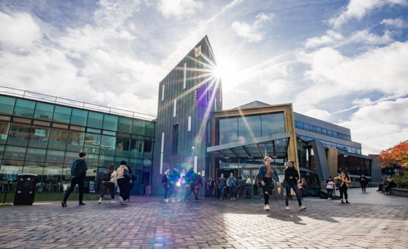 A modern building with a glass facade and a tall, narrow central structure. The sun shines brightly, creating lens flares and shadows. Several people are walking or standing near the entrance. The sky is partly cloudy, contributing to the bright atmosphere.