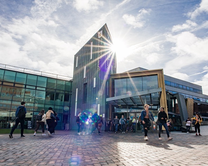 A modern building with a glass facade and a tall, narrow central structure. The sun shines brightly, creating lens flares and shadows. Several people are walking or standing near the entrance. The sky is partly cloudy, contributing to the bright atmosphere.