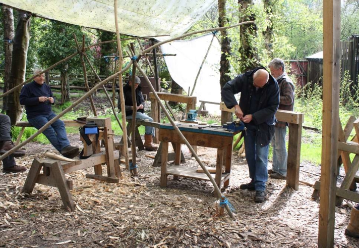 People working at wooden benches in an outdoor woodland workshop, using hand tools under a simple canopy surrounded by trees.