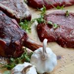 Several cuts of meat on a chopping board with bulbs of garlic and herbs.