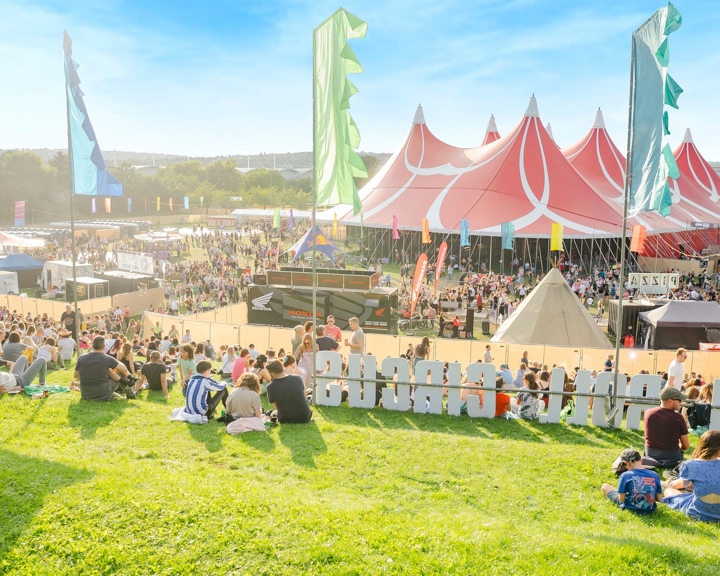 Large outdoor music festival with crowds gathered on a grassy hill and around a big red-and-white striped tent. Colorful vertical flags line the area, and smaller tents and stalls are visible in the background under a bright blue sky.