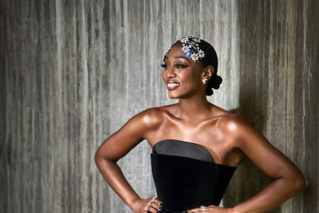 Person standing against a textured wall, wearing a strapless black dress with a structured bodice. They have short hair decorated with silver accessories and are smiling with hands on hips.