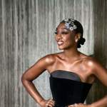 Person standing against a textured wall, wearing a strapless black dress with a structured bodice. They have short hair decorated with silver accessories and are smiling with hands on hips.