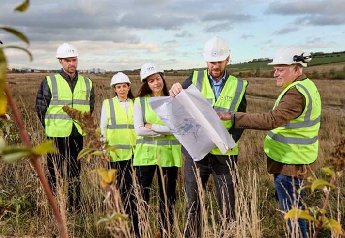 Five people in high viz jackest and hard hats stand in a field studing some plans.