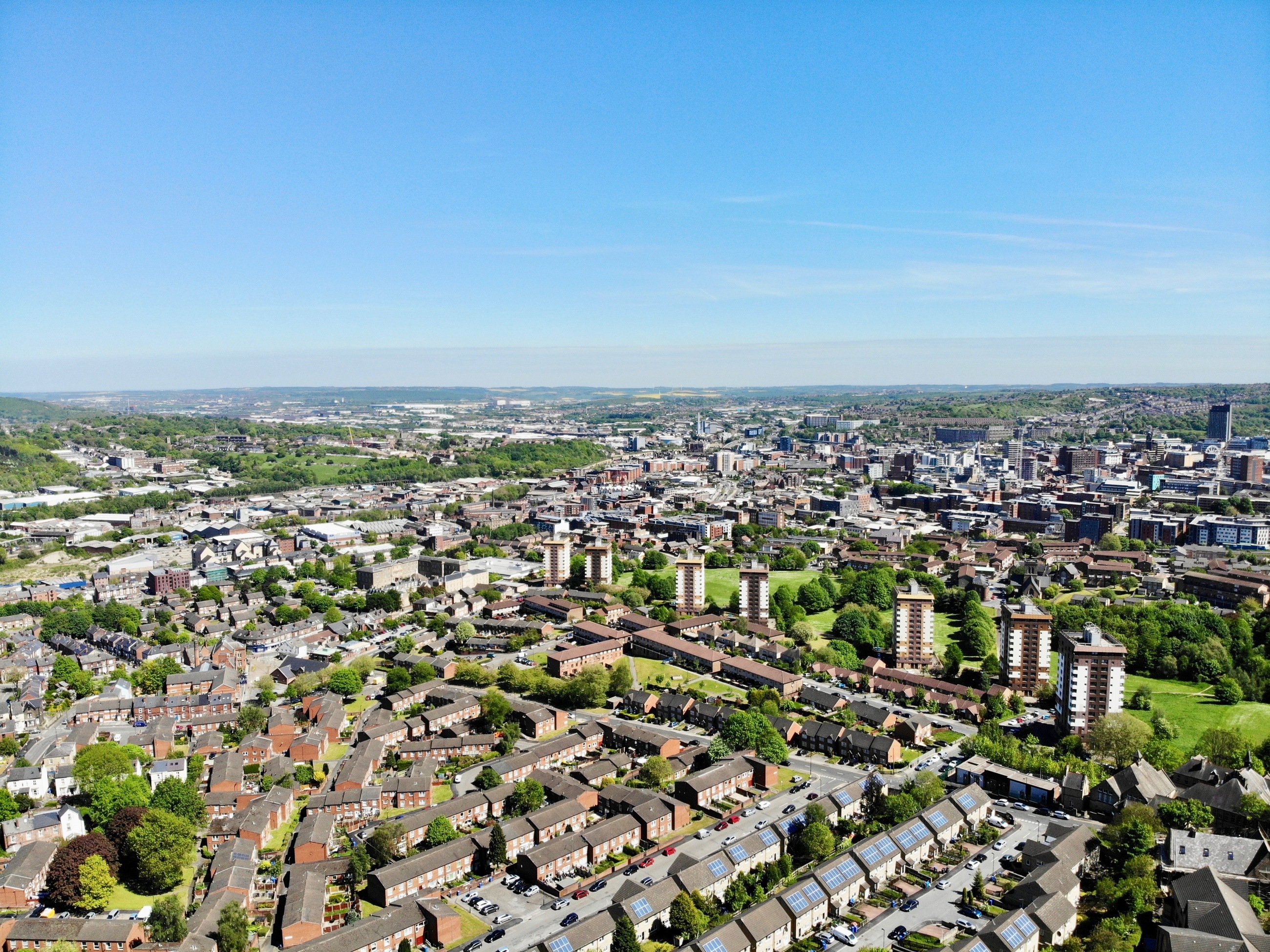 Aerial view of a city with rows of residential houses in the foreground, clusters of mid-rise apartment blocks, and green spaces scattered throughout. In the distance, the urban center features taller buildings and commercial areas, with rolling hills visible on the horizon under a clear blue sky.