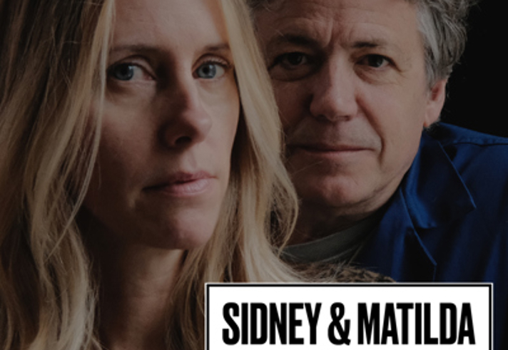 A promotional poster features two individuals shown in a close-up portrait, positioned side by side against a dark, softly blurred background. Bold white text at the top reads “XAN TYLER & DUSTY STRAY.” A boxed label in the center reads “SIDNEY & MATILDA,” with additional text below stating “APRIL 12, 7:00” and “£12.” At the bottom, small white text provides ticket information and website links, alongside a small logo for Sidney & Matilda.