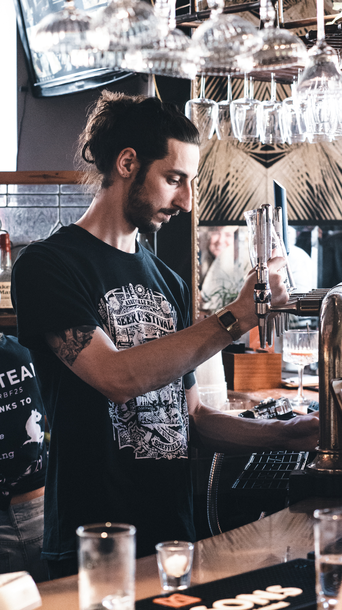 A bartender pouring a drink from a beer tap, standing behind a bar lined with glasses and bar tools, with hanging glassware overhead.