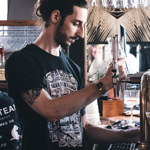 A bartender pouring a drink from a beer tap, standing behind a bar lined with glasses and bar tools, with hanging glassware overhead.