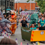 A group of participants in colorful dinosaur costumes run alongside a large yellow soapbox cart with orange chevron markings during a city street race. The cart has event branding and is surrounded by purple-and-white cones and red-and-white barriers. Crowds of spectators line both sides of the track, and a purple “Finish line” banner is visible in the background along with buildings and event signage.