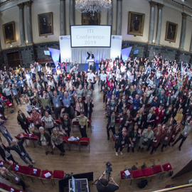 A large group of people gathered in a grand hall for the ITI Conference 2019. The hall features ornate architectural details, chandeliers, tall columns, and framed portraits on the walls. Attendees are standing and facing the stage, where a speaker is positioned at a podium beneath a screen displaying the conference name. Red chairs are arranged in rows, and a camera setup is visible in the foreground capturing the event.