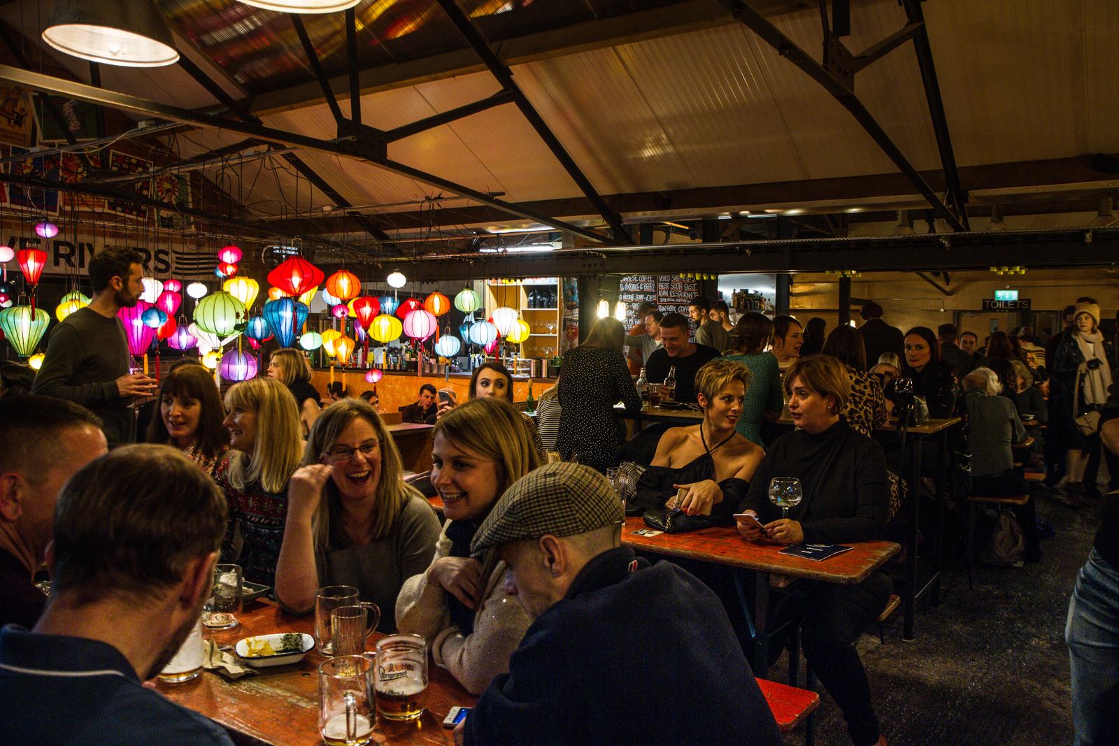 Busy indoor bar or restaurant with a lively atmosphere. The space features exposed beams and industrial-style decor, with colorful hanging lanterns creating a vibrant focal point. People are seated at wooden tables enjoying drinks and food, while others stand and converse near the bar area in the background.
