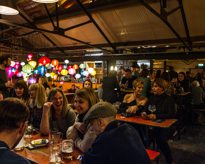 Busy indoor bar or restaurant with a lively atmosphere. The space features exposed beams and industrial-style decor, with colorful hanging lanterns creating a vibrant focal point. People are seated at wooden tables enjoying drinks and food, while others stand and converse near the bar area in the background.