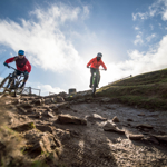 Two people mountain biking down a slope out in the countryside.