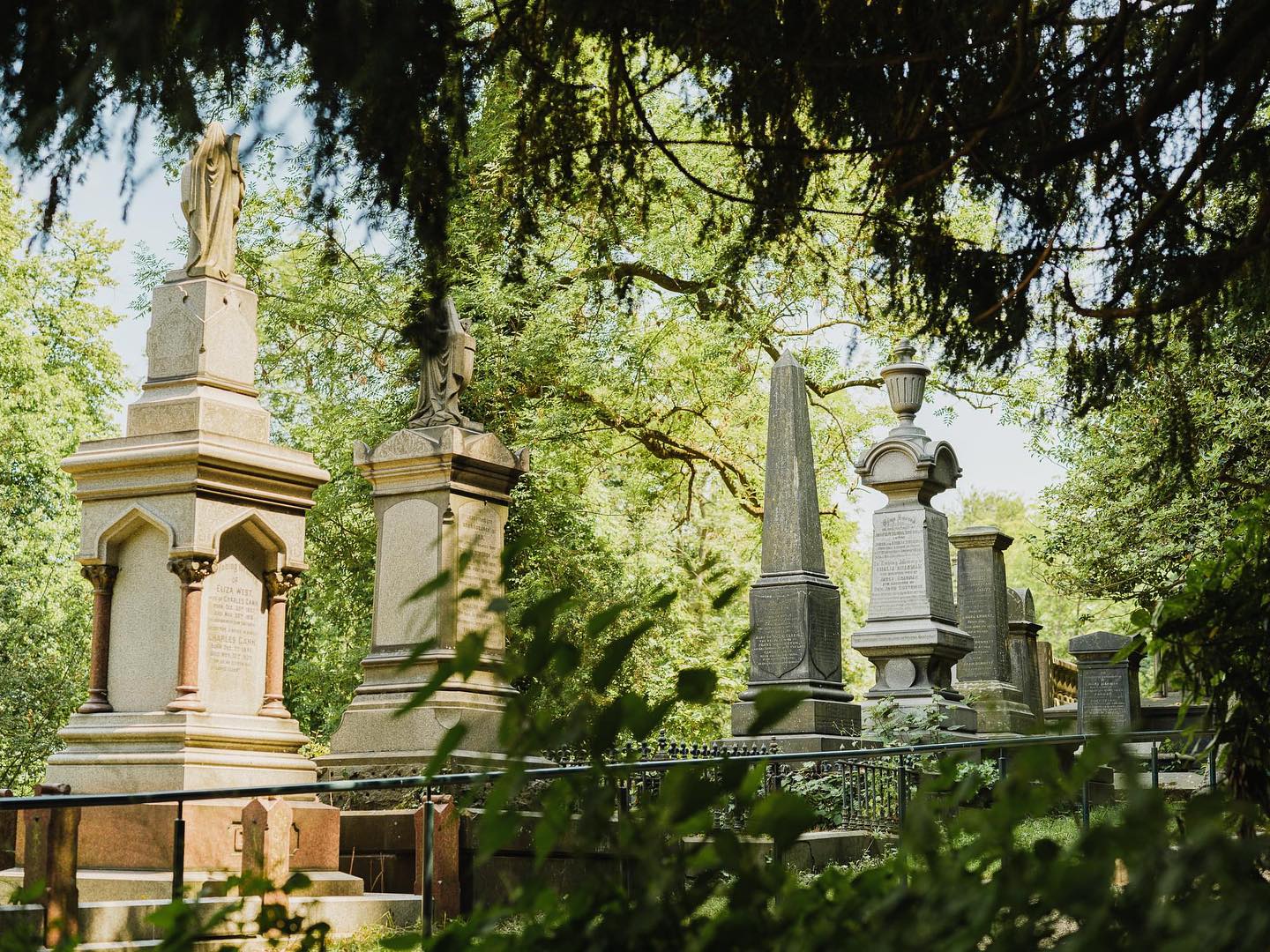 Historic cemetery scene with tall, ornate gravestones and monuments, including statues and obelisks, surrounded by lush greenery. Sunlight filters through tree branches, casting dappled light on the stone structures. Foreground features leafy plants, while large trees frame the image from above, creating a serene and shaded atmosphere.