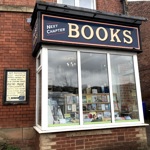 Exterior view of Next Chapter Books showing a large bay window filled with books. The shop sign above reads ‘Next Chapter BOOKS’ in cream lettering on a dark blue background, and a framed board listing subject areas is mounted on the brick wall beside the entrance.