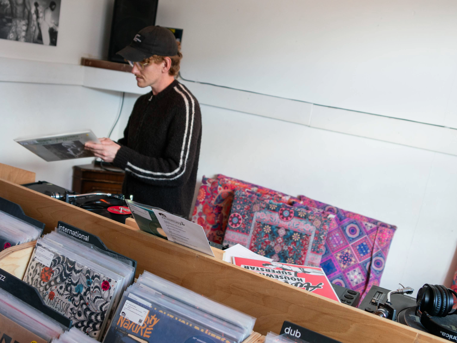 Interior of a record shop with wooden crates filled with vinyl records organised by genre, including labels such as “international” and “dub.” A person stands behind the counter holding a record near a turntable, with speakers and headphones visible. Brightly patterned fabric and posters decorate the space, creating a casual, retro atmosphere.