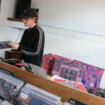 Interior of a record shop with wooden crates filled with vinyl records organised by genre, including labels such as “international” and “dub.” A person stands behind the counter holding a record near a turntable, with speakers and headphones visible. Brightly patterned fabric and posters decorate the space, creating a casual, retro atmosphere.