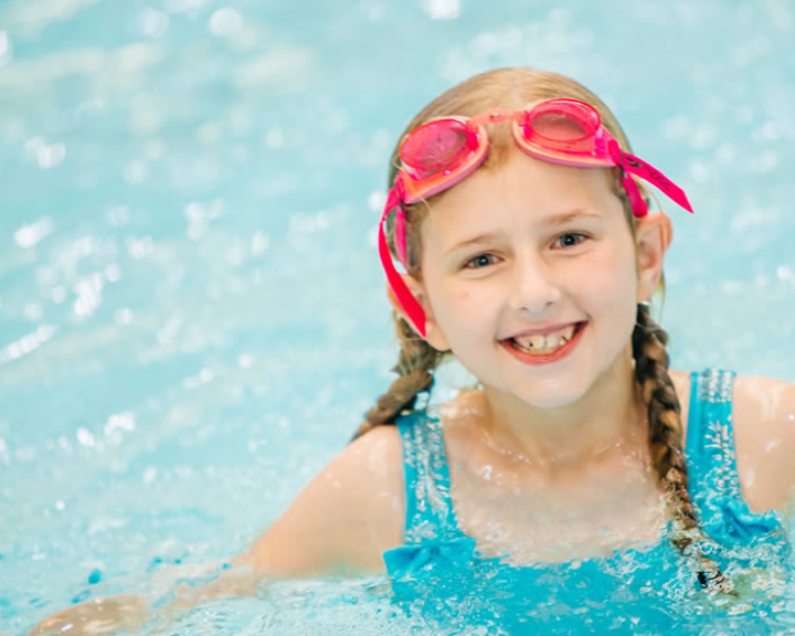 Child wearing a turquoise swimsuit and pink swimming goggles, partially submerged in a clear blue swimming pool with rippling water.