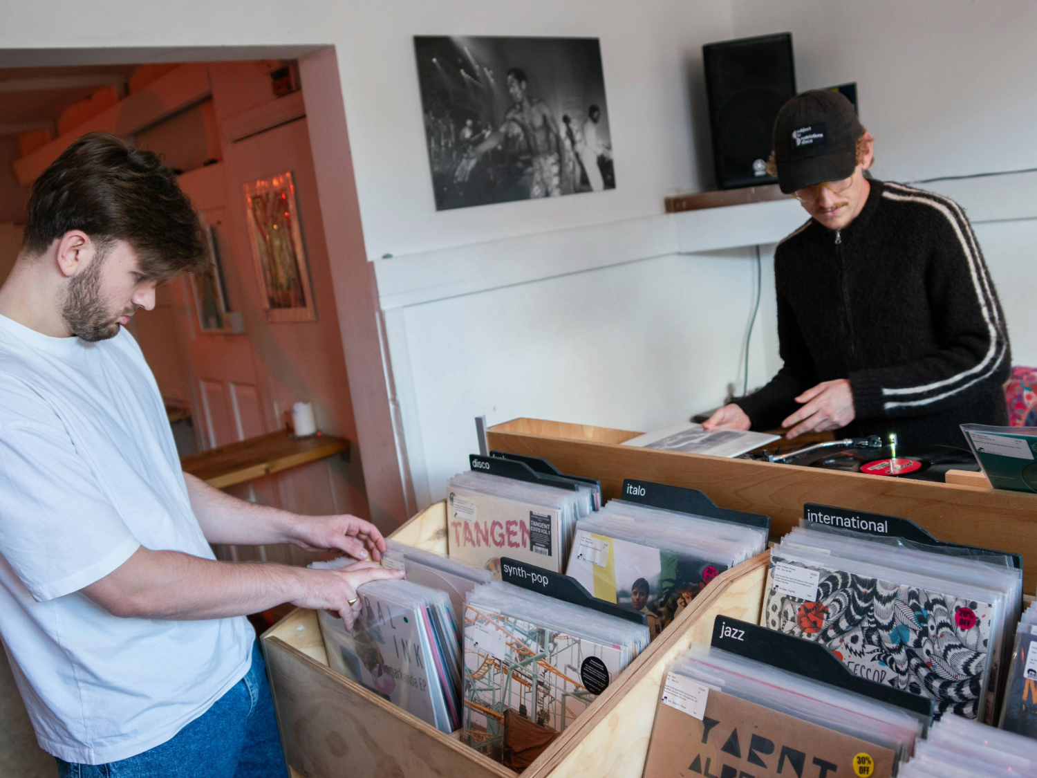 Two people in a small record shop browsing vinyl records. One person is looking through a wooden crate filled with records organised by genre, including labels such as “jazz,” “international,” and “alt.” The other person is behind a counter with more records and a speaker on the shelf. A black-and-white photo hangs on the wall, and the setting has a casual, retro feel.