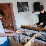 Two people in a small record shop browsing vinyl records. One person is looking through a wooden crate filled with records organised by genre, including labels such as “jazz,” “international,” and “alt.” The other person is behind a counter with more records and a speaker on the shelf. A black-and-white photo hangs on the wall, and the setting has a casual, retro feel.
