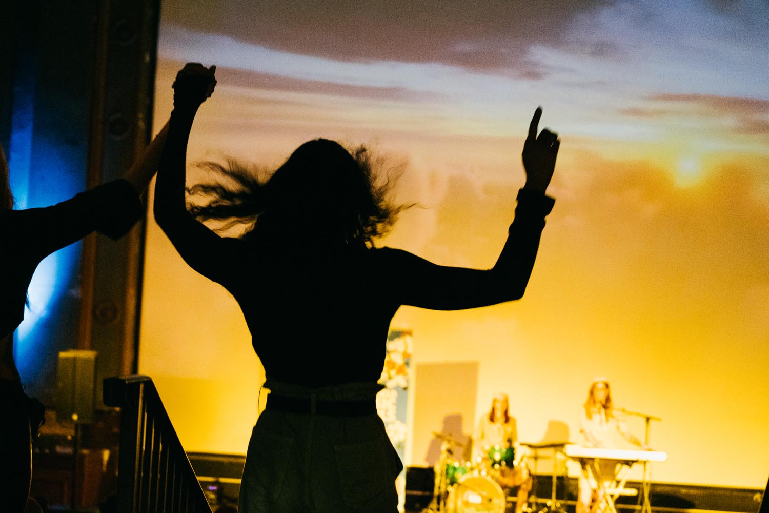 A silhouette of a person with arms raised, standing in front of a brightly lit stage where musicians are performing. The background features a large screen displaying a vivid sunset scene with orange and yellow hues. On stage, a drum set and keyboard are visible, with performers seated behind the instruments. The image captures the lively atmosphere of a concert or music event indoors.