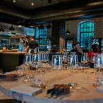 A modern, stylish restaurant interior featuring an open kitchen with a chef at work. In the foreground, a round wooden table is set with multiple empty wine glasses, folded napkins, and cutlery. The space has exposed stone walls, large arched windows, and shelves stocked with bottles. Black pendant lights hang from the ceiling, and the atmosphere is warm and contemporary.