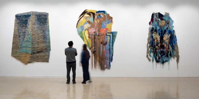 Two people standing in a gallery observing three large textile artworks mounted on a white wall. The pieces feature abstract designs with layered textures and vibrant colors, including shades of blue, green, yellow, and brown. The central artwork has long strands hanging down, creating a dynamic effect. The floor is light-colored and reflective, and the overall setting is minimalistic, emphasizing the art.