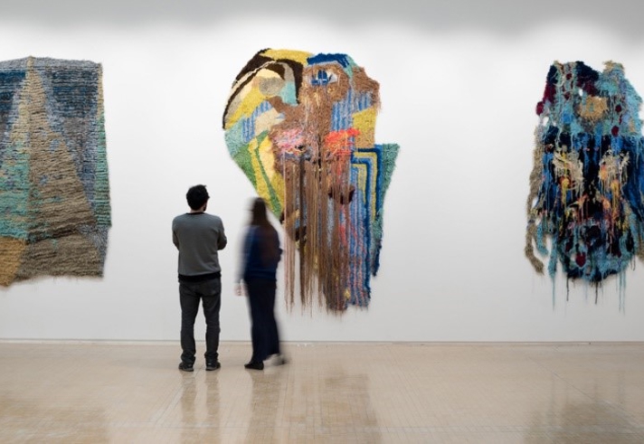 Two people standing in a gallery observing three large textile artworks mounted on a white wall. The pieces feature abstract designs with layered textures and vibrant colors, including shades of blue, green, yellow, and brown. The central artwork has long strands hanging down, creating a dynamic effect. The floor is light-colored and reflective, and the overall setting is minimalistic, emphasizing the art.