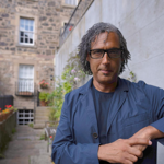 A person in a blue t-shirt and jacket leans against a brick wall.