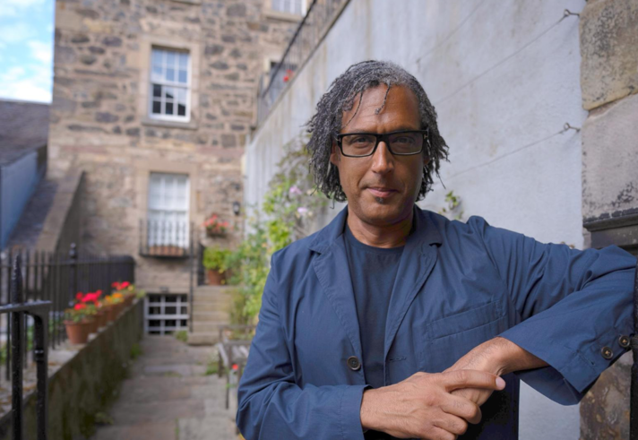 A person in a blue t-shirt and jacket leans against a brick wall.