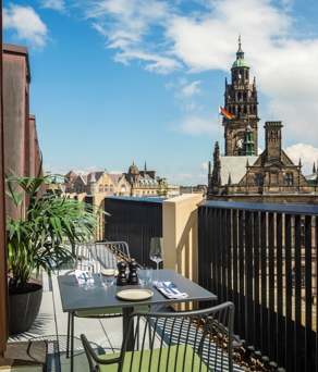 Outdoor balcony with a small dining table set for two, overlooking a historic cityscape with a tall clock tower and other ornate buildings under a partly cloudy blue sky.