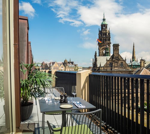 Outdoor balcony with a small dining table set for two, overlooking a historic cityscape with a tall clock tower and other ornate buildings under a partly cloudy blue sky.