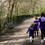 A woman and two children, all wearing Weston Park Cancer Charity Walk t-shirts, are walking away from the camera. On  their left is a row of trees and on their right is a lake.