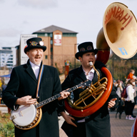 Two musicians, one playing a banjo and the other a brass sousaphone, perform at an outdoor market in Sheffield.