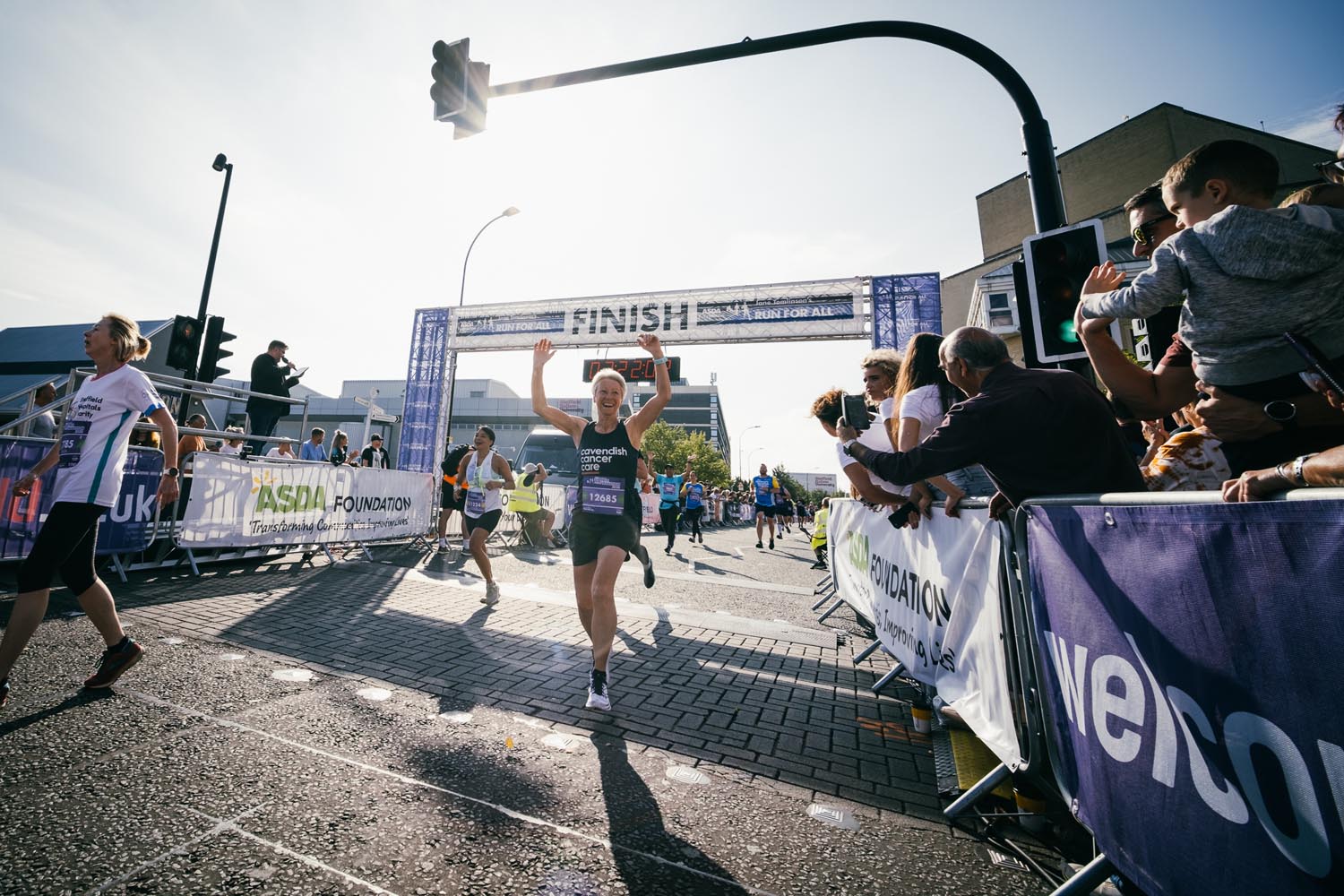 Female runner cheering as she crosses the finish line of Sheffield Half Marathon in Castlegate. The sun is shining and spectators are cheering from the right hand side of her.