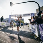 Female runner cheering as she crosses the finish line of Sheffield Half Marathon in Castlegate. The sun is shining and spectators are cheering from the right hand side of her.