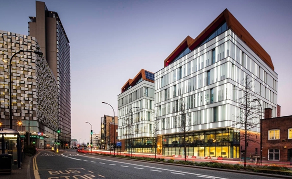 Modern city street at dusk with contemporary glass buildings featuring angular rooftops and illuminated interiors. On the left, a tall patterned building contrasts with the sleek design of the two main structures. The road in the foreground has bus lane markings, streetlights, and light trails from passing vehicles, with a clear sky in the background.