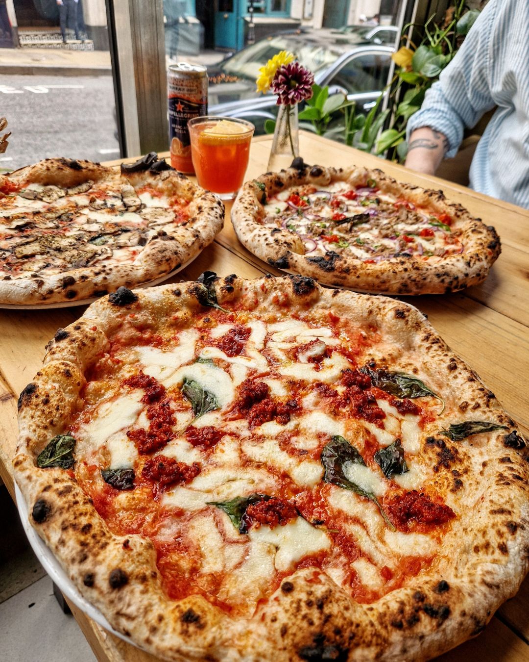 Three Neapolitan-style pizzas on a wooden table near a window, each topped with different ingredients, with a small vase of flowers and a drink beside them.