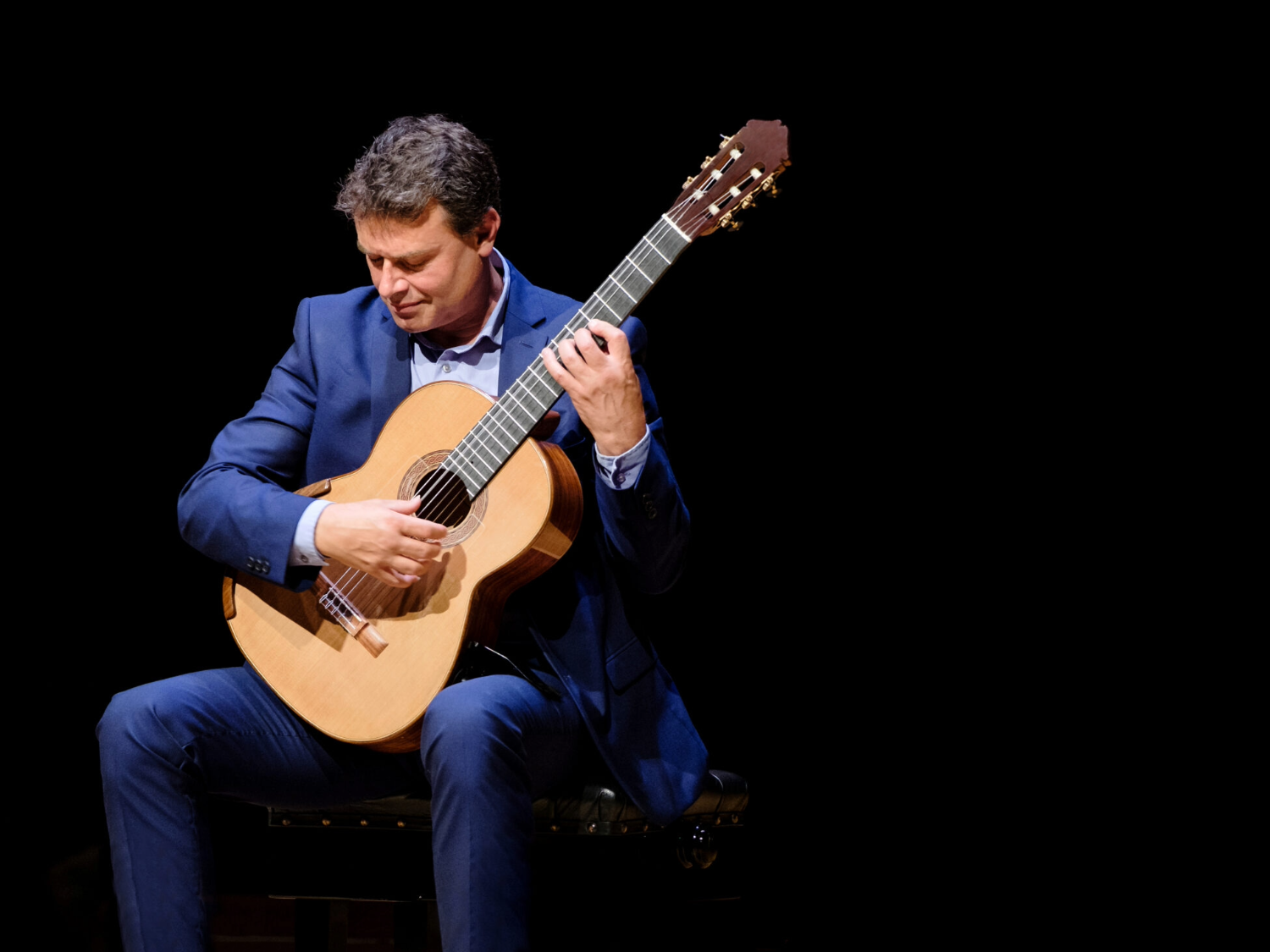 A person wearing a dark blue suit is seated on a chair, playing a classical guitar. The guitar has a light wooden body with nylon strings, and the musician’s hands are positioned on the fretboard and soundhole, suggesting a performance. The background is completely black, drawing focus to the player and the instrument.