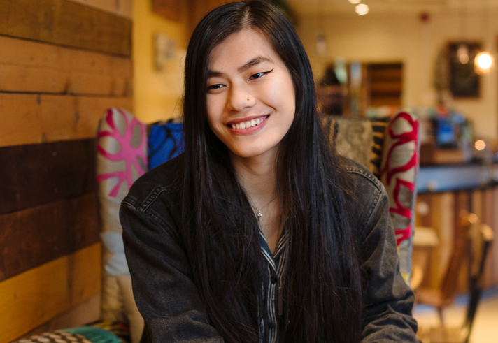 A person with long straight dark hair is seated indoors on a patterned chair. The individual is wearing a dark denim jacket over a striped shirt. The background features wooden paneling on the wall and warm ambient lighting, suggesting a cozy café or restaurant setting.