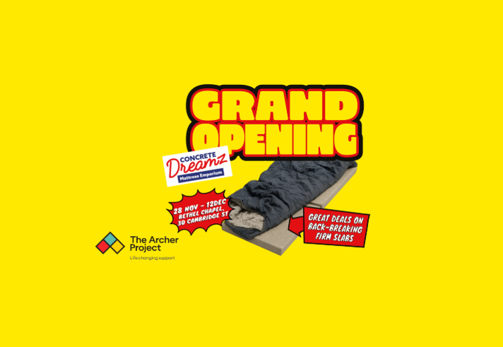 Bright yellow poster with bold red and black text reading “Grand Opening.” Below, an image of a sleeping bag on a concrete slab with a sign saying “Great deals on back-breaking firm slabs.” Another sign reads “Concrete Dreamz Mattress Emporium.” Event details: 28 Nov – 12 Dec at Bethel Chapel, 30 Cambridge St. Bottom left shows The Archer Project logo with tagline “Life changing support.”
