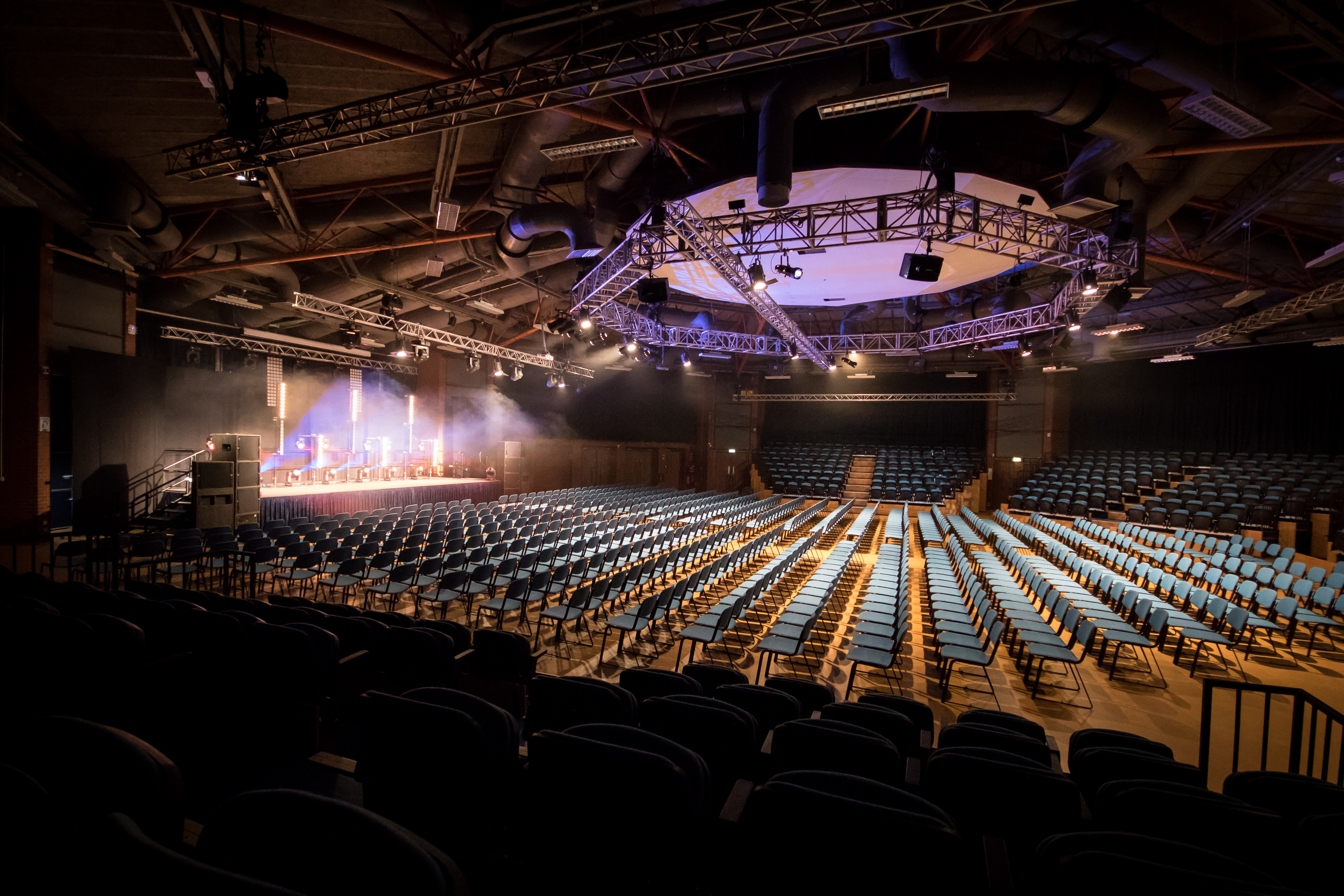 Large auditorium set up in theatre style with rows of blue chairs facing a stage. The stage is illuminated with bright spotlights and features podiums and equipment. Overhead, a circular truss with lighting rigs hangs from the exposed industrial ceiling with beams and ducts. The space is dimly lit except for the stage area, creating a dramatic atmosphere.
