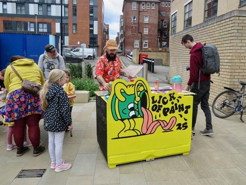 Outdoor street scene with a small art stall featuring a bright yellow counter painted with a cartoon character and the words ‘LICK OF PAINT 25.’ Several people, including children, are gathered around the stall. The background shows modern brick buildings, a bicycle leaning against a wall, and a paved walkway.