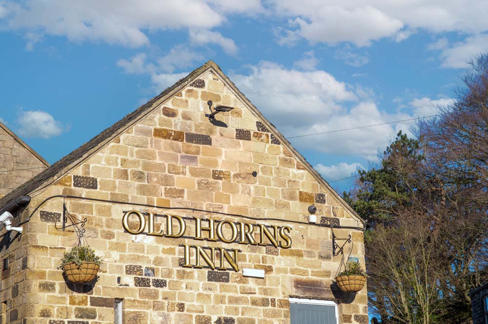 The gable end of the Old Horns Inn, featuring its stone façade, hanging flower baskets and gold lettering, set against a bright blue sky with scattered clouds.