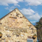 The gable end of the Old Horns Inn, featuring its stone façade, hanging flower baskets and gold lettering, set against a bright blue sky with scattered clouds.