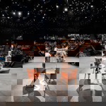 Two speakers standing on a stage in front of a full audience inside a theatre with star‑like ceiling lights.