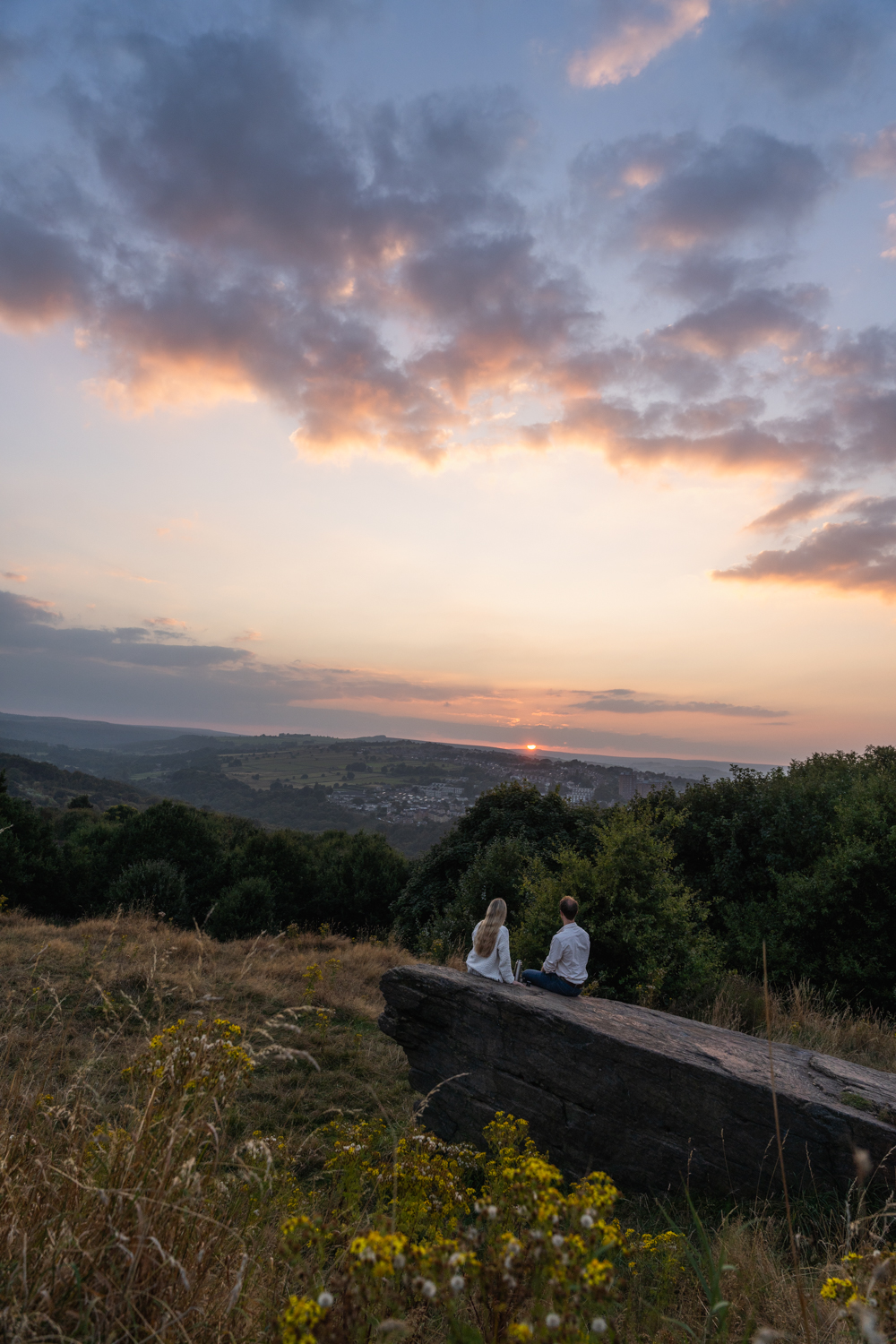 Two people sitting on a large flat rock in a grassy hillside area, overlooking a valley with scattered trees and distant houses. The sky is filled with soft clouds illuminated by the warm colours of a sunset, casting a serene glow across the landscape. Yellow wildflowers are visible in the foreground.