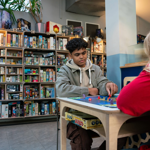 Two people are sat at a table, in a cafe, playing a board game and drinking coffees. Behind them is shelving stacked full of all different board games.