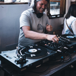 A DJ operating turntables and a mixer inside a bright room, with plants and a decorative mirror in the background.