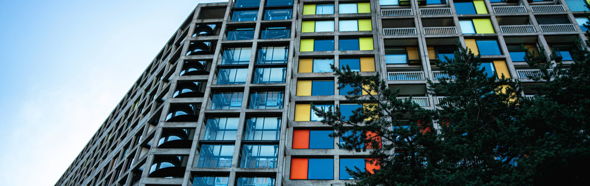 A brutalist high-rise apartment block made of concrete and glass. The windows are interspersed with bright panels in yellows and oranges.